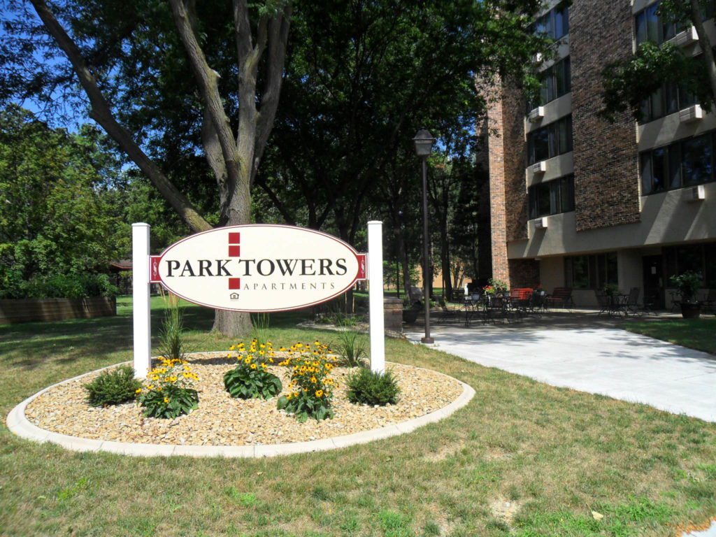 A large sign reading Park Towers Apartments stands in a landscaped area with yellow flowers, in front of a multi-story apartment building surrounded by trees and greenery.