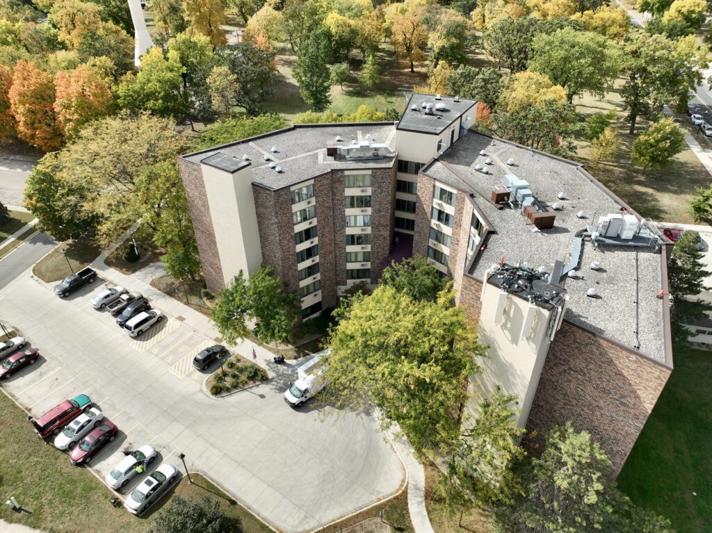Aerial view of a mid-rise apartment building surrounded by trees with autumn foliage. Cars are parked in a lot alongside the building, and a road curves around the property.