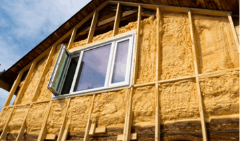 A house exterior wall under construction with exposed yellow spray foam insulation between wooden framing; a white window is installed, and the sky is visible above.
