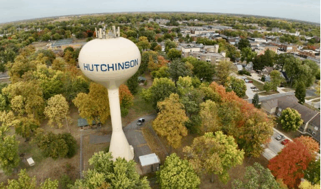A large water tower labeled HUTCHINSON stands among trees with fall foliage, surrounded by buildings and houses in a suburban area under a partly cloudy sky.