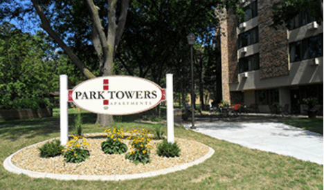 A sign reading Park Towers Apartments stands on a landscaped area with small plants and flowers in front of an apartment building surrounded by trees on a sunny day.