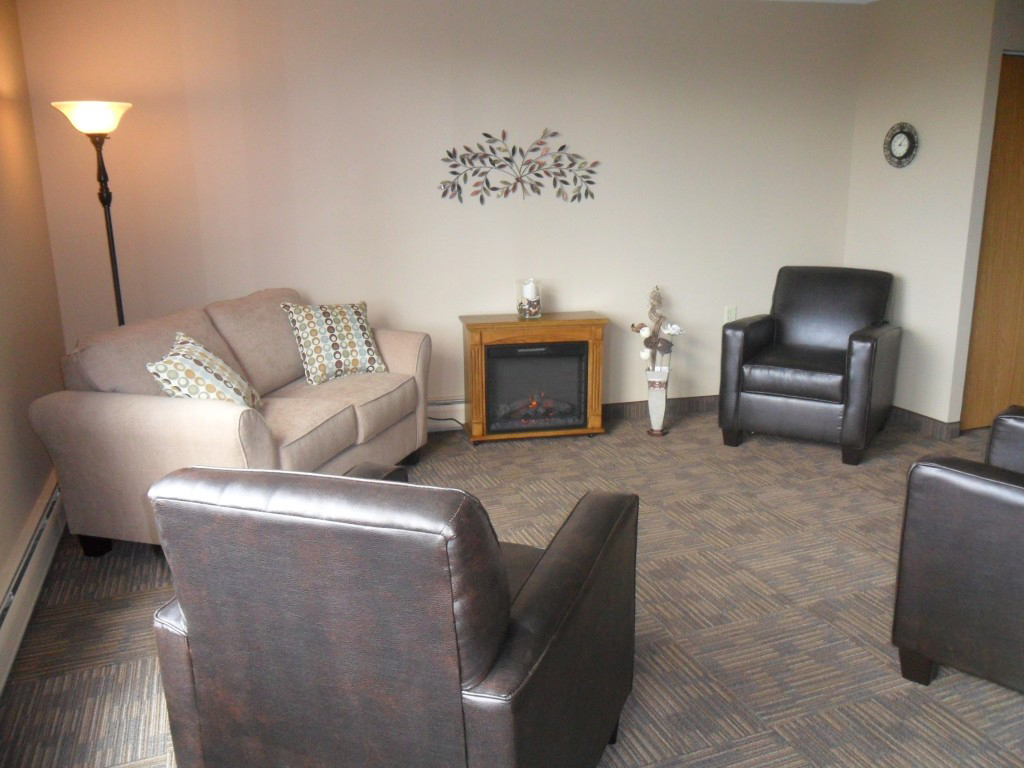 A cozy living room with a beige sofa, two dark brown armchairs, a floor lamp, a small electric fireplace, decorative pillows, wall art, and neutral-colored carpet and walls.