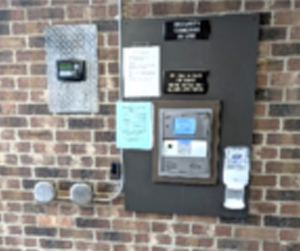 A wall with a brick background featuring various mounted devices, including an electronic meter, a thermostat, notices, and a hand sanitizer dispenser.
